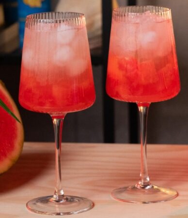 Dwa kieliszki różowego mocktailu z lodem, obok kawałka arbuza i puszki wody kokosowej z guaraną i magnezem QF Quality Food
