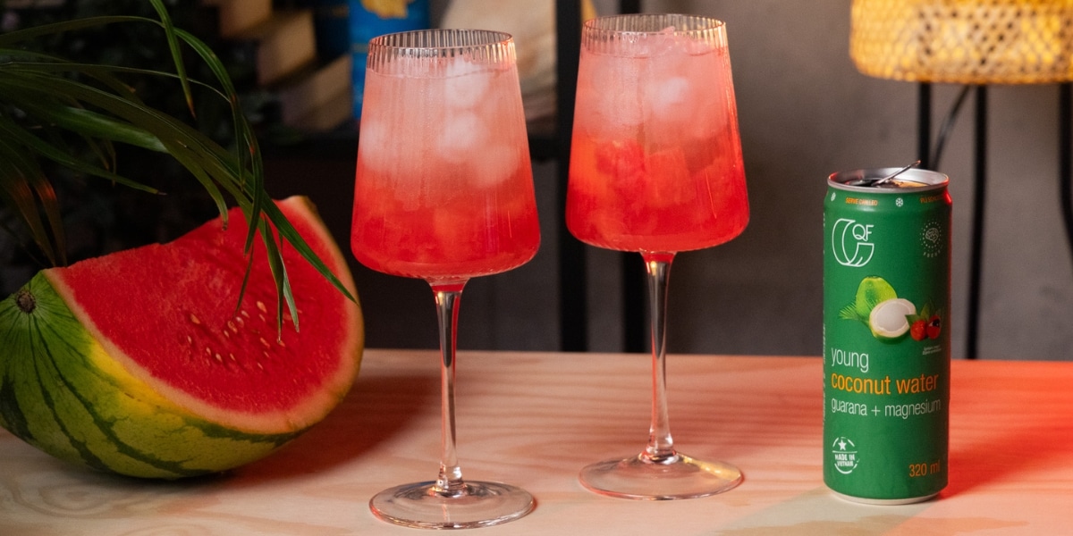 Dwa kieliszki różowego mocktailu z lodem, obok kawałka arbuza i puszki wody kokosowej z guaraną i magnezem QF Quality Food