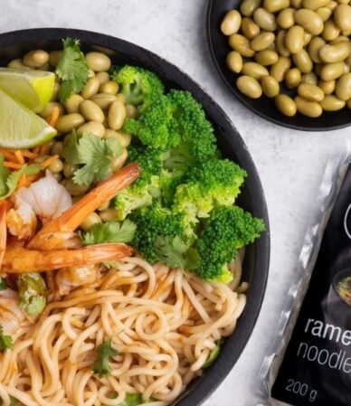 Czarna miska z krewetkami, ramenem, edamame, brokułem, marchewką i limonką, obok opakowanie makaronu proteinowego Ramen QF Active