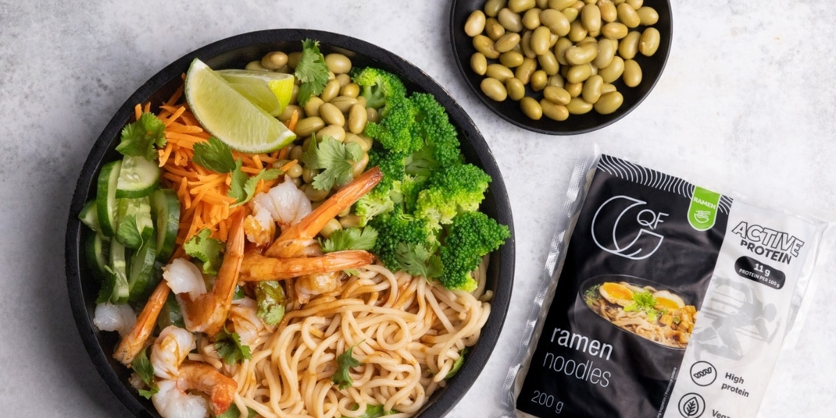 Czarna miska z krewetkami, ramenem, edamame, brokułem, marchewką i limonką, obok opakowanie makaronu proteinowego Ramen QF Active