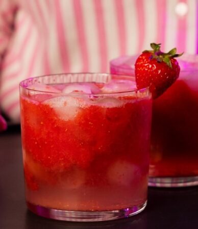 Szklanka czerwonego mocktailu truskawkowego z lodem i truskawką na brzegu, obok puszki wody kokosowej z limonką QF Quality Food
