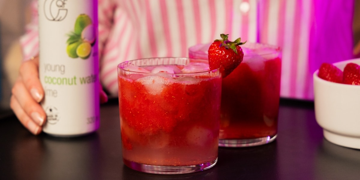 Szklanka czerwonego mocktailu truskawkowego z lodem i truskawką na brzegu, obok puszki wody kokosowej z limonką QF Quality Food