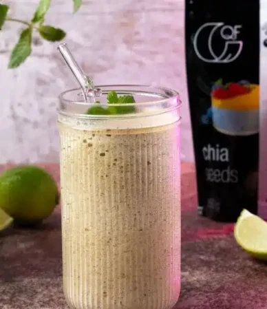 Un smoothie crémeux à la menthe servi dans un grand verre côtelé avec une paille, accompagné de quartiers de citron vert, d'une canette d'eau de coco QF et d'un sachet de graines de chia.