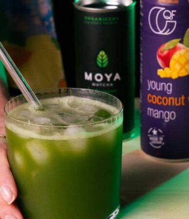 Matcha congelado con hielo y una pajita en un vaso sostenido en las manos, junto a latas de agua de coco QF Mango y Moya Matcha.